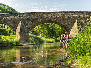Zwei Wanderer werfen Steine in den Bach unter der historischen Steinbergsbrücke in Dernau im Ahrtal.
