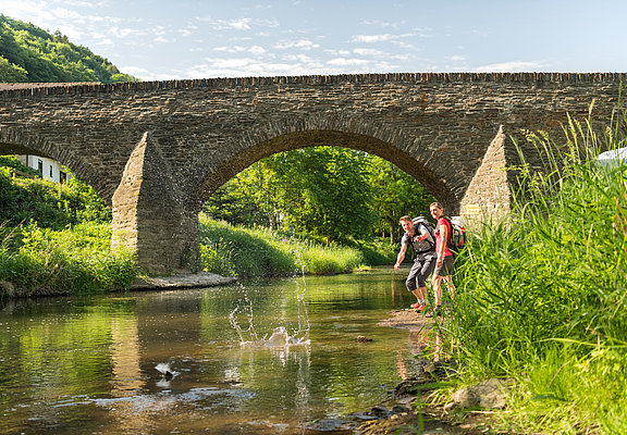Zwei Wanderer werfen Steine in den Bach unter der historischen Steinbergsbrücke in Dernau im Ahrtal.