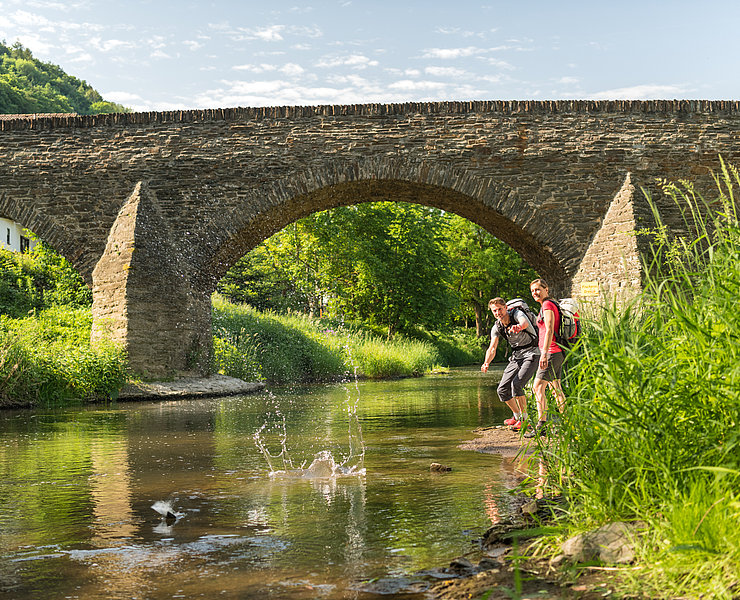 Zwei Wanderer werfen Steine in den Bach unter der historischen Steinbergsbrücke in Dernau im Ahrtal.